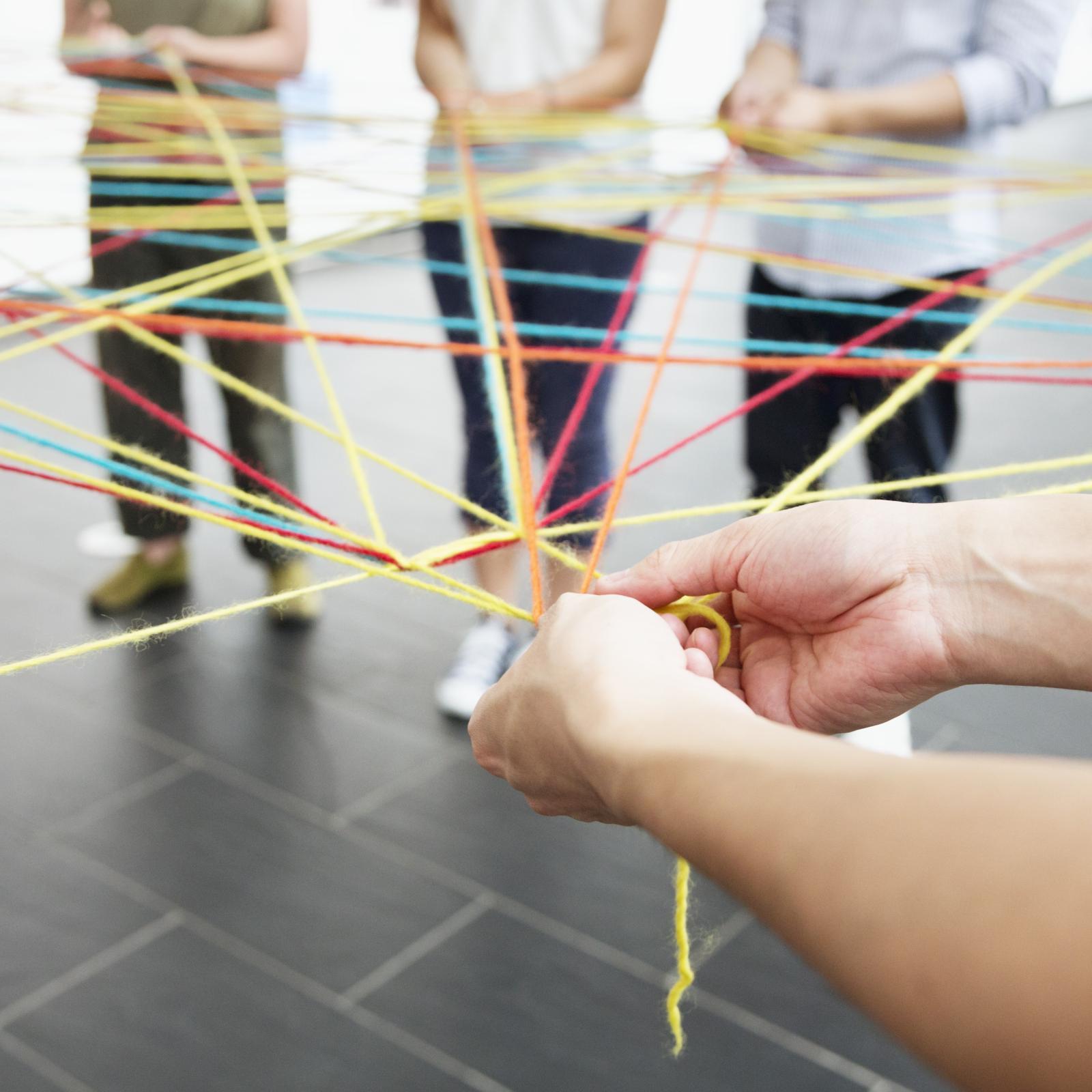 Foto von einer Gruppe weißer Personen, die im Kreis stehen und mehrere bunte Wollfäden halten, die netzwerkartig zwischen ihnen entlang führen.