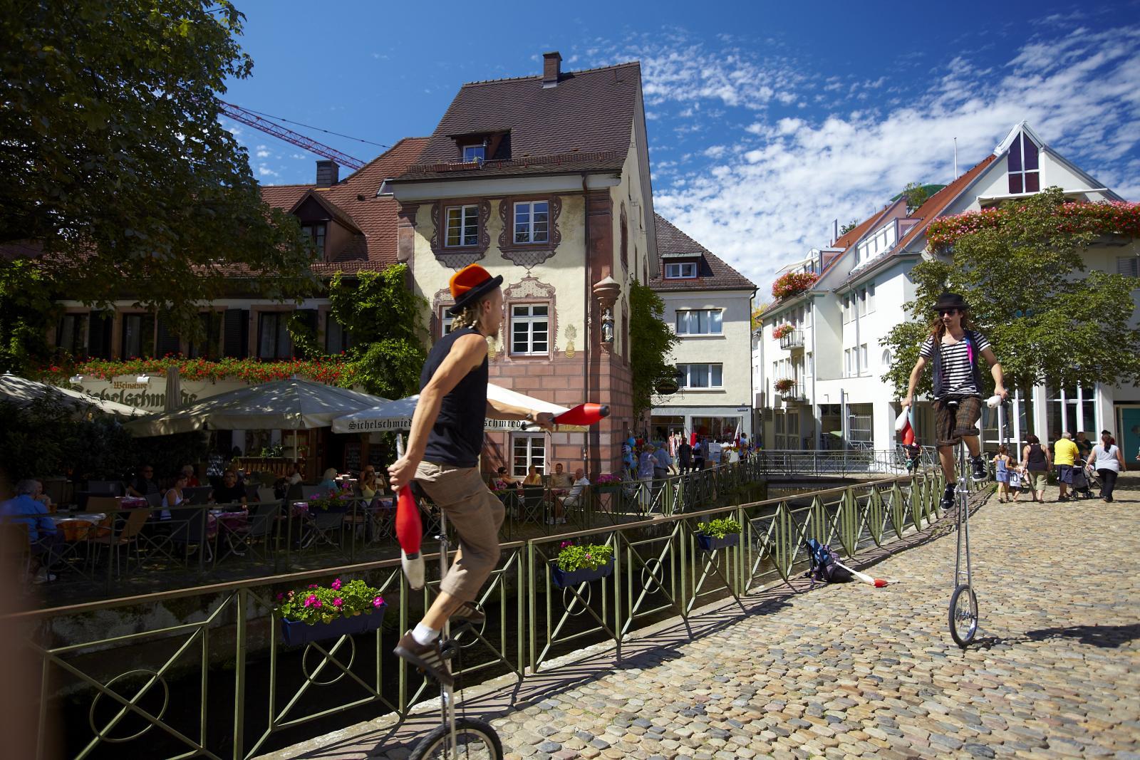 Eine Straße in der Altstadt von Freiburg. Zwei Einradfahrer fahren über eine Brücke, im Hintergrund sind die schmalen und niedrigen Gebäude der Altstadt zu sehen.