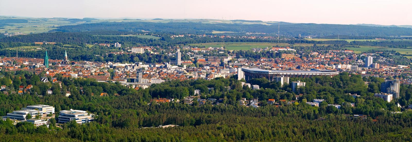 Blick über Kaiserslautern: Die Stadt ist umgeben von grüner Landschaft. Kirchtürme, Hochhäuser und ein Stadion ragen zwischen niedrigeren rot gedeckten Dächern hervor.