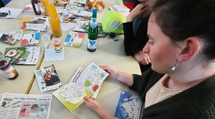 Das Foto zeigt eine weiße Frau mit dunklen Haaren und Ohrschmuck, die an einem Tisch sitzt, auf dem viele bunte Papiere und Flyer liegen sowie Getränkeflaschen stehen. 