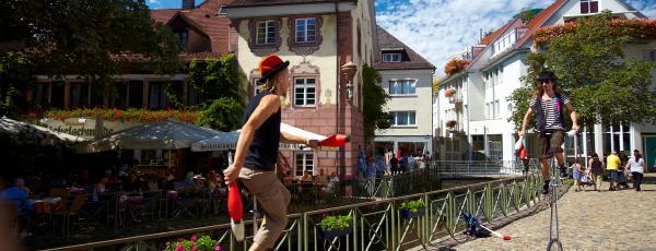 Eine Straße in der Altstadt von Freiburg. Zwei Einradfahrer fahren über eine Brücke, im Hintergrund sind die schmalen und niedrigen Gebäude der Altstadt zu sehen.