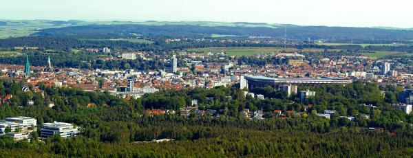Blick über Kaiserslautern: Die Stadt ist umgeben von grüner Landschaft. Kirchtürme, Hochhäuser und ein Stadion ragen zwischen niedrigeren rot gedeckten Dächern hervor.